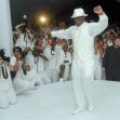 A crowd of photographers and attendees watches as a man in white attire dances at an event. A crowd of photographers and attendees watches as a man in white attire dances at an event.