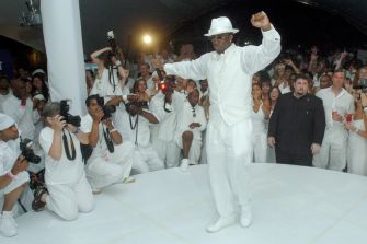 A crowd of photographers and attendees watches as a man in white attire dances at an event.