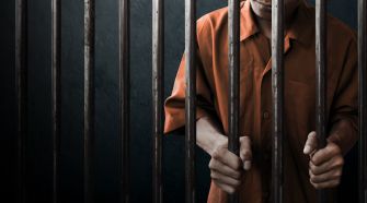 A person in an orange prison jumpsuit gripping metal bars inside a prison cell.