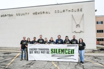 A group of Greenpeace supporters standing outside the Morton County Memorial Courthouse, holding a banner that reads "ENERGY TRANSFER: WE WILL NOT BE SILENCED."