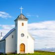A small white church with a cross on the roof against a blue sky and mountains in the background. A small white church with a cross on the roof against a blue sky and mountains in the background.