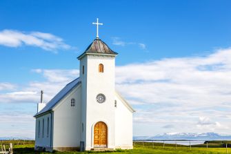 A small white church with a cross on the roof against a blue sky and mountains in the background.