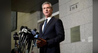 California Governor Gavin Newsom speaking at a press conference with multiple microphones in front of him.