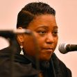 A woman speaking into a microphone at a public event, appearing serious and focused. A woman speaking into a microphone at a public event, appearing serious and focused.