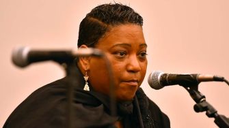 A woman speaking into a microphone at a public event, appearing serious and focused.