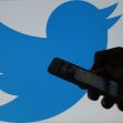 Silhouette of a hand holding a smartphone in front of a large Twitter logo. Silhouette of a hand holding a smartphone in front of a large Twitter logo.