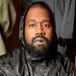 Image of Kanye West wearing a black hooded jacket, seated against a backdrop of blurred clothing items. Image of Kanye West wearing a black hooded jacket, seated against a backdrop of blurred clothing items.