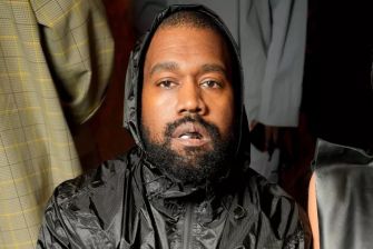 Image of Kanye West wearing a black hooded jacket, seated against a backdrop of blurred clothing items.