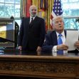 A man sitting at a desk with papers in hand, while another man stands beside him, both in an office setting adorned with flags and official emblems. A man sitting at a desk with papers in hand, while another man stands beside him, both in an office setting adorned with flags and official emblems.