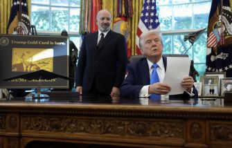 A man sitting at a desk with papers in hand, while another man stands beside him, both in an office setting adorned with flags and official emblems.