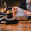 A gavel is placed on a wooden desk with blurred background of legal books and a person holding papers, symbolizing a court proceeding. A gavel is placed on a wooden desk with blurred background of legal books and a person holding papers, symbolizing a court proceeding.
