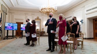 Participants, including President Biden and First Lady Jill Biden, standing with hands over their hearts during an event in the White House, with a video screen in the background.