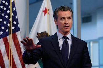 California Governor Gavin Newsom speaking at a press conference with American and California flags in the background.
