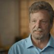 A man with short hair and a beard, wearing a blue shirt, speaks in an interview setting. A man with short hair and a beard, wearing a blue shirt, speaks in an interview setting.