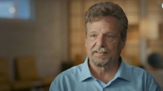 A man with short hair and a beard, wearing a blue shirt, speaks in an interview setting.