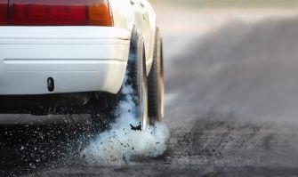 A close-up view of a white car's rear tire leaving smoke and debris on a racing surface during a drag race.