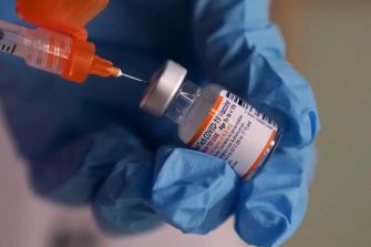 Close-up of a gloved hand holding a COVID-19 vaccine vial with a syringe poised to draw the vaccine.