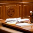 A courtroom setting featuring a wooden bench with the scales of justice emblem and a pile of documents on the table. A courtroom setting featuring a wooden bench with the scales of justice emblem and a pile of documents on the table.