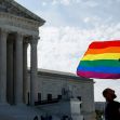 A person holding a flag featuring the rainbow plus trans pride colors in front of the Supreme Court building. A person holding a flag featuring the rainbow plus trans pride colors in front of the Supreme Court building.