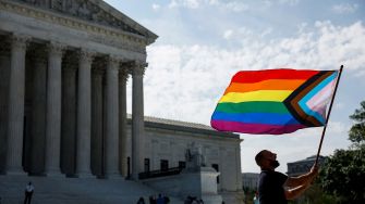 A person holding a flag featuring the rainbow plus trans pride colors in front of the Supreme Court building.