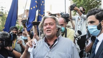 Steve Bannon surrounded by media outside a public event, gesturing and speaking while wearing a mask.