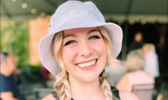 A smiling young woman wearing a white bucket hat, with braids, in a casual outdoor setting.