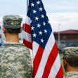 Two soldiers in military uniforms stand with an American flag at a public event. Two soldiers in military uniforms stand with an American flag at a public event.
