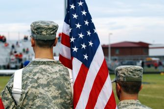 Two soldiers in military uniforms stand with an American flag at a public event.