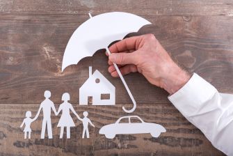 A hand holding a paper umbrella over cut-out figures of a family, a house, and a car, symbolizing insurance protection.