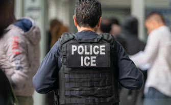 A law enforcement officer wearing an "ICE" vest is seen in a crowded area.