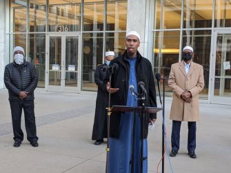 A group of men, including a speaker in traditional attire, addresses the media outside a courthouse, following the conviction of militia leader Michael Hari for a mosque bombing.
