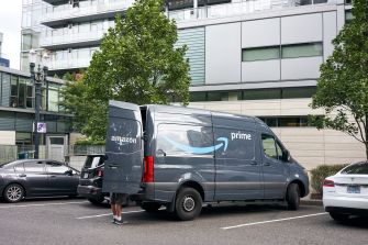 Amazon delivery van parked in a lot, with the back door open, surrounded by trees and nearby buildings.