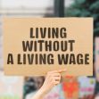 A person holding a sign that reads "LIVING WITHOUT A LIVING WAGE" at a protest or demonstration. A person holding a sign that reads "LIVING WITHOUT A LIVING WAGE" at a protest or demonstration.