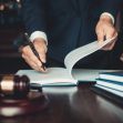 A person in a suit writes in a legal document with a gavel and scales of justice visible on a desk. A person in a suit writes in a legal document with a gavel and scales of justice visible on a desk.