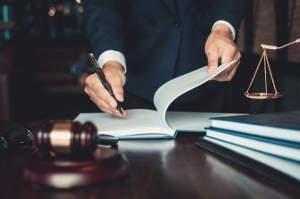 A person in a suit writes in a legal document with a gavel and scales of justice visible on a desk.