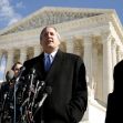 A man in a suit speaks at a press conference in front of the Supreme Court building, flanked by supporters and microphones. A man in a suit speaks at a press conference in front of the Supreme Court building, flanked by supporters and microphones.