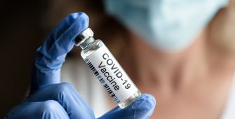 A person in a blue glove holds a vial labeled "COVID-19 Vaccine" while wearing a face mask.