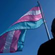 A silhouette of a person standing in front of a waving transgender pride flag against a blue sky. A silhouette of a person standing in front of a waving transgender pride flag against a blue sky.