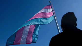 A silhouette of a person standing in front of a waving transgender pride flag against a blue sky.