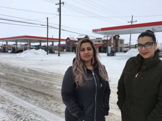 Two women standing outside in a snowy area near a gas station.