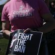 A person wearing a shirt that says "Combat Veteran" holds a sign that reads "FIGHT THE BAN." A person wearing a shirt that says "Combat Veteran" holds a sign that reads "FIGHT THE BAN."