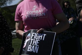 A person wearing a shirt that says "Combat Veteran" holds a sign that reads "FIGHT THE BAN."