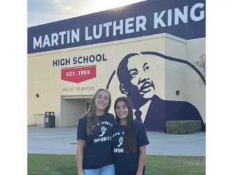 Two female athletes from Martin Luther King High School standing in front of the school building, wearing t-shirts that promote girls' sports.