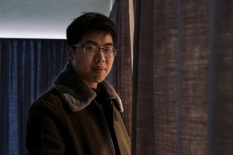 A young man standing indoors, wearing glasses and a coat, with curtains in the background.