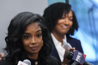 A focused press conference featuring a woman speaking into a microphone, with another woman smiling in the background.