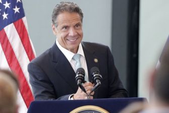 Former New York Governor Andrew Cuomo speaking at an event, with an American flag in the background.