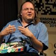 A man with long brown hair and glasses speaking while gesturing, sitting in front of a backdrop related to personal democracy and Google. A man with long brown hair and glasses speaking while gesturing, sitting in front of a backdrop related to personal democracy and Google.