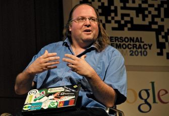 A man with long brown hair and glasses speaking while gesturing, sitting in front of a backdrop related to personal democracy and Google.