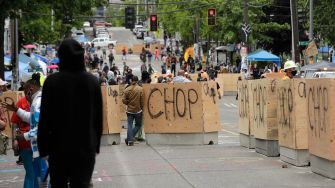 A view of barricades labeled "CHOP" during the Capitol Hill Occupied Protest zone in Seattle, with protesters and vehicles in the background.