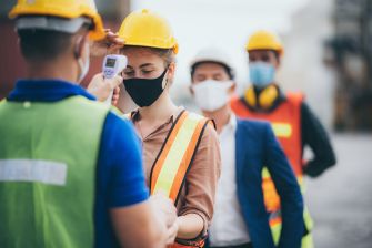 Workers wearing safety gear and masks are being screened for temperature at a workplace, highlighting health measures during the COVID-19 pandemic.